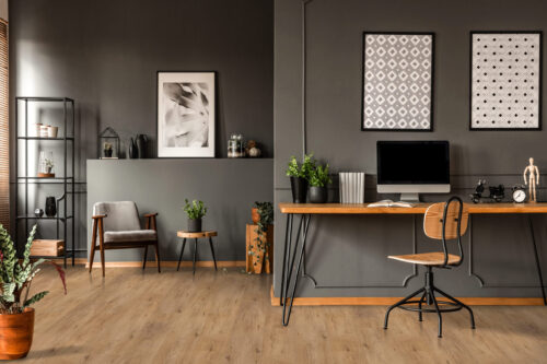 Modern home office with rustic hardwood flooring in a warm brown tone, featuring a minimalist wooden desk, vintage chair, and dark accent walls. Premium hardwood floors from Rustic Wood Floors Supply.