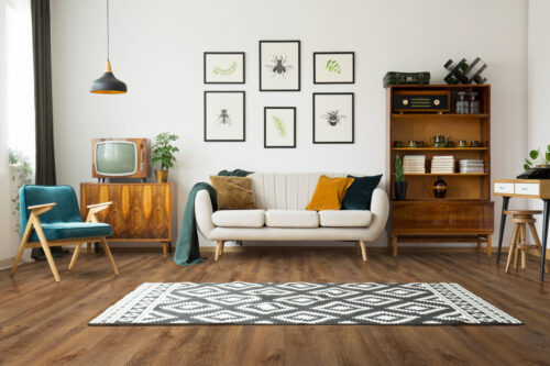 Rustic wide plank hardwood flooring in a warm brown tone, showcasing a living room interior with mid-century modern furniture, a patterned rug, and white walls. Ideal for demonstrating the aesthetic of installing rustic wood floors from Rustic Wood Floors Supply