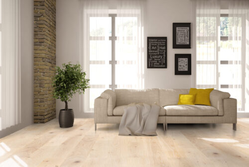 Bright and airy living room with wide-plank light hardwood flooring. The natural wood grain adds warmth to the modern decor, featuring a neutral-toned sofa, accent pillows, and brick wall detail. Premium hardwood flooring from Rustic Wood Floors Supply.