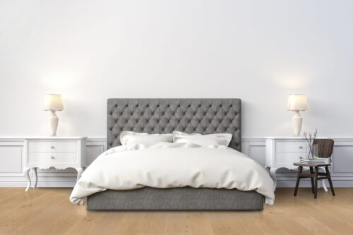 Light-toned, natural wood grain hardwood flooring from Rustic Wood Floors Supply in a bedroom setting, showcasing a grey tufted bed, white bedding, and matching white nightstands, highlighting the flooring's warm and inviting aesthetic