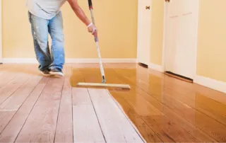 A person applying a glossy finish to a wooden floor with a roller, wearing jeans and gloves, in a room with light yellow walls and a white door.