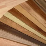 A fan-shaped display of various wooden flooring samples in different shades and grain patterns, arranged on a light wood floor background.