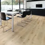 A wide-angle shot of a modern, open-plan living and dining room with large windows. The floor is covered in light-colored engineered hardwood flooring. A dining table with black chairs is in the foreground and a black couch and entertainment center are in the background.
