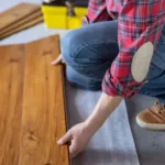A contractor in a red plaid shirt and jeans is kneeling on the floor, installing a plank of pre-finished hardwood flooring.