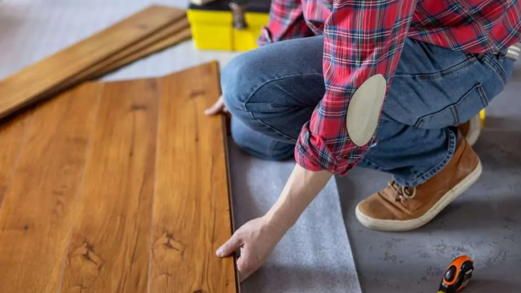 A contractor in a red plaid shirt and jeans is kneeling on the floor, installing a plank of pre-finished hardwood flooring.