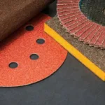 How to Choose the Right Sandpaper Grit and Sealers When Refinishing Floors? A close-up still life arrangement of various sanding materials and abrasives on a dark background. The items include a rolled sheet of brown sandpaper, a circular red sanding disc with vacuum holes, a stack of angled flap discs for an angle grinder, a rectangular sanding sponge, and a cylindrical sanding brush wheel.